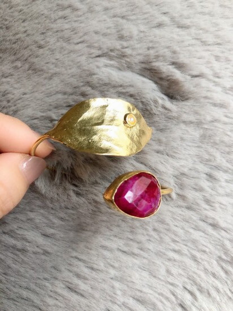 May include: Two gold rings. One ring is a leaf shape with a small, round, clear stone. The other ring has a teardrop-shaped, deep red stone. Both rings have a gold band. The rings are displayed on a gray, textured surface.