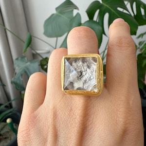May include: A gold-toned square ring with a clear, textured center. The ring is worn on a finger, showcasing the intricate details of the design. The background features green foliage.