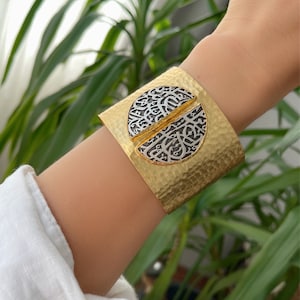 Peut inclure: Un bracelet manchette en or avec une finition martelée et un motif circulaire argenté orné de calligraphie arabe complexe.