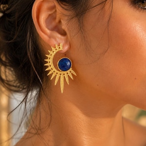 May include: A gold-toned earring featuring a semi-circular sunburst design with radiating pointed rays. A circular, faceted deep blue stone is set within the center of the crescent shape, creating a contrast between the metallic gold and blue.