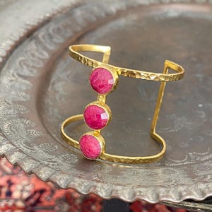 May include: Gold bangle bracelet with three pink gemstone accents. The bracelet has a hammered texture.