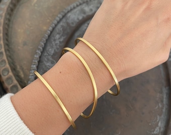 May include: Three gold-toned metal bangles stacked on a person's wrist. The bangles are simple and have a smooth, polished finish.