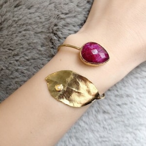 May include: A gold-toned bracelet featuring a large, faceted, ruby-colored gemstone and a gold leaf with a small pearl accent. The bracelet is worn on a wrist, with a soft, gray background.