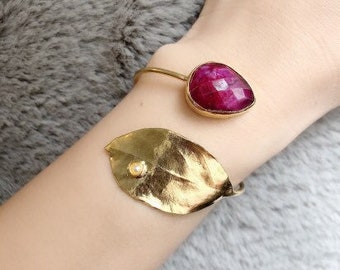 Könnte beinhalten: Ein goldfarbenes Armband mit einem großen, facettierten, rubinroten Edelstein und einem goldenen Blatt mit einem kleinen Perlenakzent. Das Armband wird am Handgelenk getragen, vor einem weichen, grauen Hintergrund.