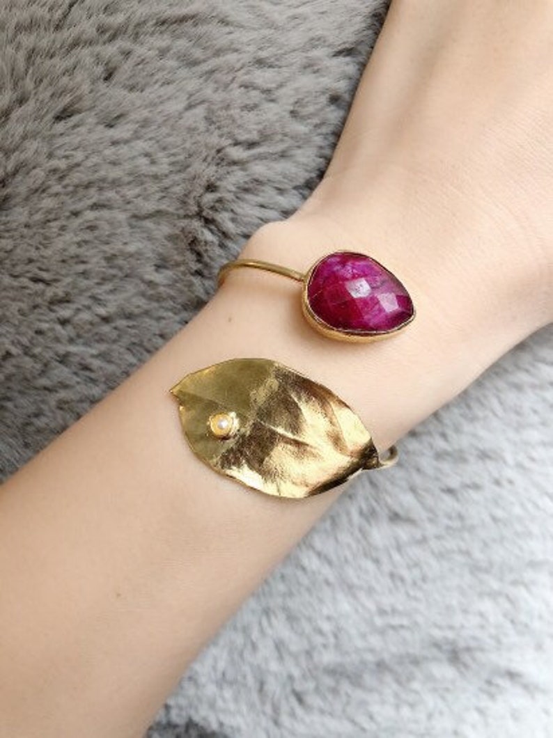 May include: A gold-toned bracelet featuring a large, faceted, ruby-colored gemstone and a gold leaf with a small pearl accent. The bracelet is worn on a wrist, with a soft, gray background.