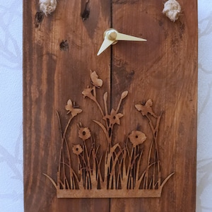 May include: A wooden wall clock with a rustic brown finish. The clock face features a laser-cut design of flowers and butterflies. The clock hands are made of gold metal.