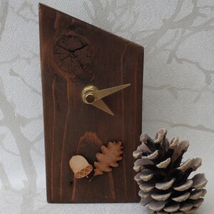 May include: A wooden clock with a brown stain and a gold-coloured hand. The clock is shaped like a triangle and has an acorn and oak leaf design. A pine cone is in the foreground.