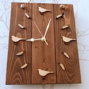 May include: A rustic wooden clock with a brown finish. The clock face is surrounded by small wooden birds and feathers. The clock hands are white.