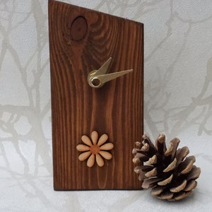 May include: A wooden clock with a brown stain and a simple design. The clock has a single hand and a small wooden flower design. The clock is sitting on a white surface next to a pine cone.