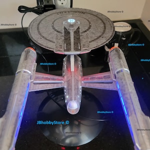 K&ouml;nnte beinhalten: Detailliertes Modell des Star Trek Raumschiffs USS Enterprise NCC-1701-D, mit einem grauen Untertassenteil und zwei Gondeln mit blauer und roter Beleuchtung. Das Modell steht auf einer schwarzen Oberfl&auml;che, mit einem Netzkabel.