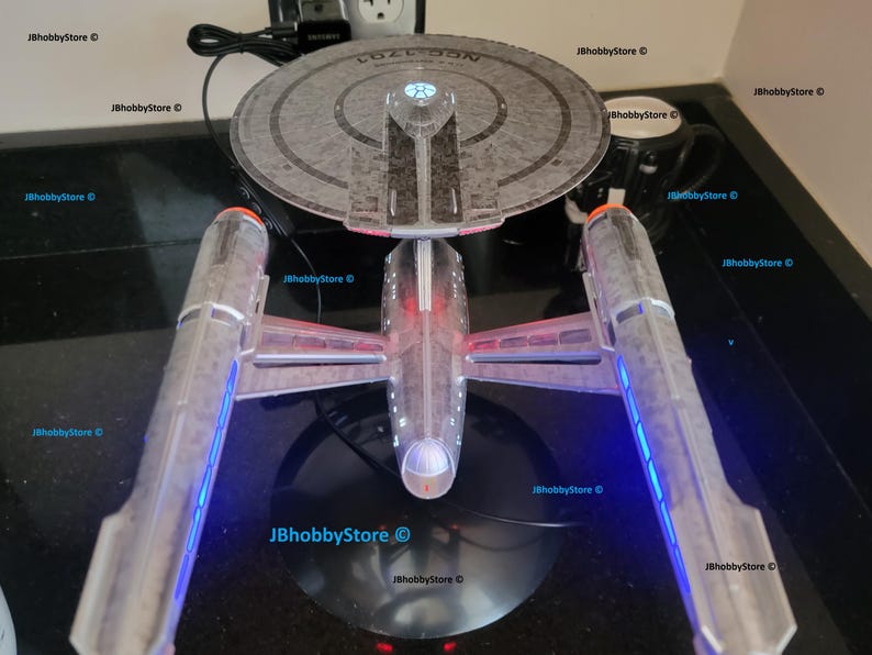 K&ouml;nnte beinhalten: Detailliertes Modell des Star Trek Raumschiffs USS Enterprise NCC-1701-D, mit einem grauen Untertassenteil und zwei Gondeln mit blauer und roter Beleuchtung. Das Modell steht auf einer schwarzen Oberfl&auml;che, mit einem Netzkabel.