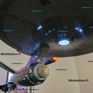 K&ouml;nnte beinhalten: Ein detailliertes Modell des Raumschiffs Enterprise mit beleuchteten Lichtern und aufwendigen Details. Das Modell zeigt den ikonischen Untertassenabschnitt des Schiffs mit dem Text "NCC-1701". Das Modell hat eine metallische Oberfl&auml;che mit orangefarbenen und blauen Lichtern.