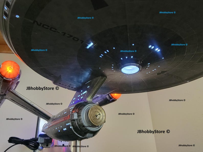 K&ouml;nnte beinhalten: Ein detailliertes Modell des Raumschiffs Enterprise mit beleuchteten Lichtern und aufwendigen Details. Das Modell zeigt den ikonischen Untertassenabschnitt des Schiffs mit dem Text "NCC-1701". Das Modell hat eine metallische Oberfl&auml;che mit orangefarbenen und blauen Lichtern.