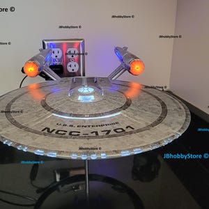 K&ouml;nnte beinhalten: Ein detailliertes Modell der U.S.S. Enterprise NCC-1701 aus Star Trek. Das Modell hat ein silbernes und graues Farbschema mit beleuchteten Lichtern, einschlie&szlig;lich orangefarbener Triebwerk-Gondeln und blauen Akzenten. Das Modell wird auf einem St&auml;nder pr&auml;sentiert.