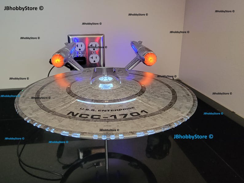 K&ouml;nnte beinhalten: Ein detailliertes Modell der U.S.S. Enterprise NCC-1701 aus Star Trek. Das Modell hat ein silbernes und graues Farbschema mit beleuchteten Lichtern, einschlie&szlig;lich orangefarbener Triebwerk-Gondeln und blauen Akzenten. Das Modell wird auf einem St&auml;nder pr&auml;sentiert.
