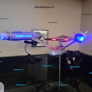 K&ouml;nnte beinhalten: Ein Modell des Raumschiffs Enterprise mit blauen und roten Lichtern. Das Modell ist silber und grau, mit einem St&auml;nder. Das Bild enth&auml;lt auch ein schwarzes Netzkabel und ein schwarzes Objekt mit einem wei&szlig;en Gesicht.