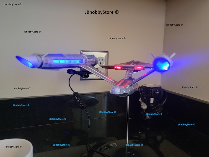 K&ouml;nnte beinhalten: Ein Modell des Raumschiffs Enterprise mit blauen und roten Lichtern. Das Modell ist silber und grau, mit einem St&auml;nder. Das Bild enth&auml;lt auch ein schwarzes Netzkabel und ein schwarzes Objekt mit einem wei&szlig;en Gesicht.