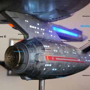 K&ouml;nnte beinhalten: Detailliertes Modell eines Raumschiffs, grau mit roten und wei&szlig;en Akzenten. Das Modell hat beleuchtete Fenster und einen blau leuchtenden Bereich. Das Schiff ist auf einem St&auml;nder montiert und zeigt sein komplexes Design.