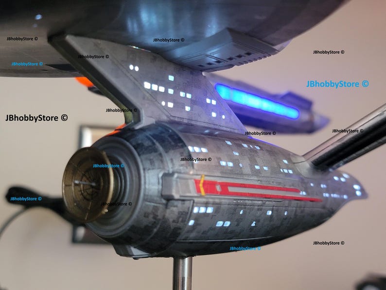 K&ouml;nnte beinhalten: Detailliertes Modell eines Raumschiffs, grau mit roten und wei&szlig;en Akzenten. Das Modell hat beleuchtete Fenster und einen blau leuchtenden Bereich. Das Schiff ist auf einem St&auml;nder montiert und zeigt sein komplexes Design.