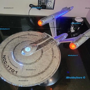 K&ouml;nnte beinhalten: Ein detailliertes Modell der U.S.S. Enterprise NCC-1701, einem Raumschiff aus dem Star Trek-Franchise. Das Modell verf&uuml;gt &uuml;ber beleuchtete Lichter, darunter blaue und orangefarbene, und ist vor einem dunklen Hintergrund platziert. Das Modell ist ein Sammlerst&uuml;ck.