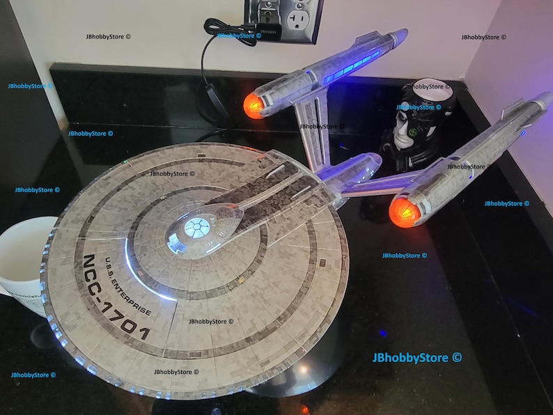 K&ouml;nnte beinhalten: Ein detailliertes Modell der U.S.S. Enterprise NCC-1701, einem Raumschiff aus dem Star Trek-Franchise. Das Modell verf&uuml;gt &uuml;ber beleuchtete Lichter, darunter blaue und orangefarbene, und ist vor einem dunklen Hintergrund platziert. Das Modell ist ein Sammlerst&uuml;ck.