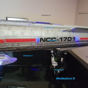 K&ouml;nnte beinhalten: Ein Modell des Raumschiffs Enterprise mit dem Text "NCC-1701" und roten und gelben Akzenten. Das Schiff hat eine silberne und graue Farbgebung mit blauer Beleuchtung. Das Bild stammt von JBhobbyStore.