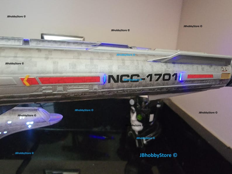K&ouml;nnte beinhalten: Ein Modell des Raumschiffs Enterprise mit dem Text "NCC-1701" und roten und gelben Akzenten. Das Schiff hat eine silberne und graue Farbgebung mit blauer Beleuchtung. Das Bild stammt von JBhobbyStore.