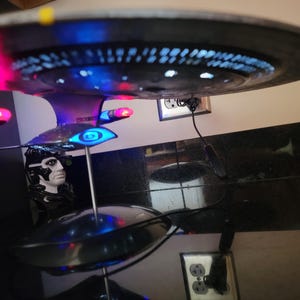 May include: A model of a starship with a black hull and illuminated blue and pink lights. The model is mounted on a stand, with a bust of a character from the Star Trek series in the foreground. The ship's design is detailed.