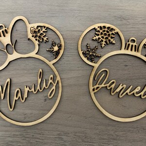 May include: Two wooden Christmas ornaments shaped like Mickey Mouse ears with a bow on one and a snowflake design on both. The ornaments are personalized with the names "Marluz" and "Daniele".