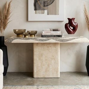 May include: A stone console table with a textured top and central pedestal base, featuring three amber glass bowls, a stack of books, and a deep red abstract sculpture. Flanking the table are two tall black vases filled with dried pampas grass.