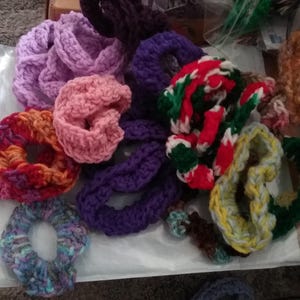 May include: A collection of colorful crocheted hair scrunchies in various patterns and colors, including purple, pink, green, red, yellow, and blue.