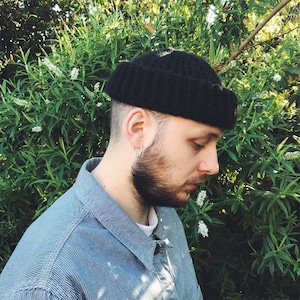 May include: A person wearing a black knitted beanie with a ribbed texture. The beanie is pulled down low on their forehead. The person is wearing a blue denim shirt with a white undershirt.