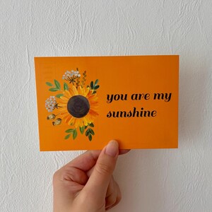 May include: A rectangular orange postcard featuring a painted sunflower with green leaves and small white flowers on the left side. The text you are my sunshine is printed in black cursive lettering on the right side of the orange card surface.