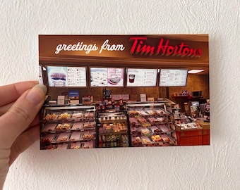 May include: A postcard with the text "greetings from Tim Hortons" on a brown background. The image shows the interior of a Tim Hortons restaurant with a display case full of donuts and other baked goods.