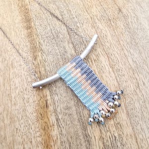 Puede incluir: Un collar de cadena de plata con un colgante tejido en colores azul, gris y melocotón. El colgante tiene una barra de plata en la parte superior y cuentas de plata en la parte inferior.