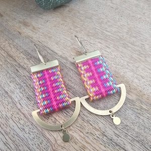 Può includere: Un paio di orecchini dorati con accenti in tessuto intrecciato rosa, blu e giallo. Gli orecchini presentano un design a semicerchio con un piccolo ciondolo rotondo.