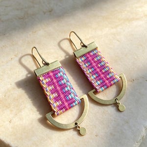 May include: A pair of drop earrings featuring a rectangular woven fabric section in vibrant shades of magenta, light blue, and yellow. Each earring is attached to a brass hook and finished with a brass semi-circular base and a small circular charm.