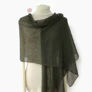 May include: An olive green shawl draped over a cream-colored turtleneck sweater. The shawl has a delicate, open-weave pattern and hangs loosely, creating a flowing silhouette. A small pink flower is pinned near the neckline. The shawl appears soft and lightweight.