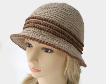 Puede incluir: Un sombrero cloche de ganchillo beige con un ala pequeña. El sombrero presenta dos bandas marrones alrededor de la corona y un borde festoneado en el ala. Este sombrero hecho a mano es un accesorio elegante.