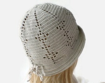 May include: A crocheted hat with a light beige color and a delicate openwork pattern. The hat has a wide brim and a bow at the back.