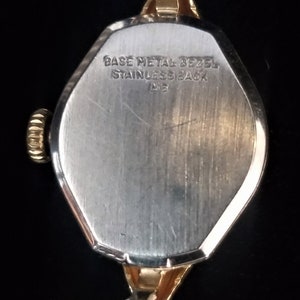 K&ouml;nnte beinhalten: Die R&uuml;ckseite einer goldfarbenen Armbanduhr mit einem silberfarbenen Geh&auml;use. Das Geh&auml;use hat einen erh&ouml;hten Rand und der Text "BASE METAL BEZEL STAINLESS BACK M15" ist in das Geh&auml;use graviert.