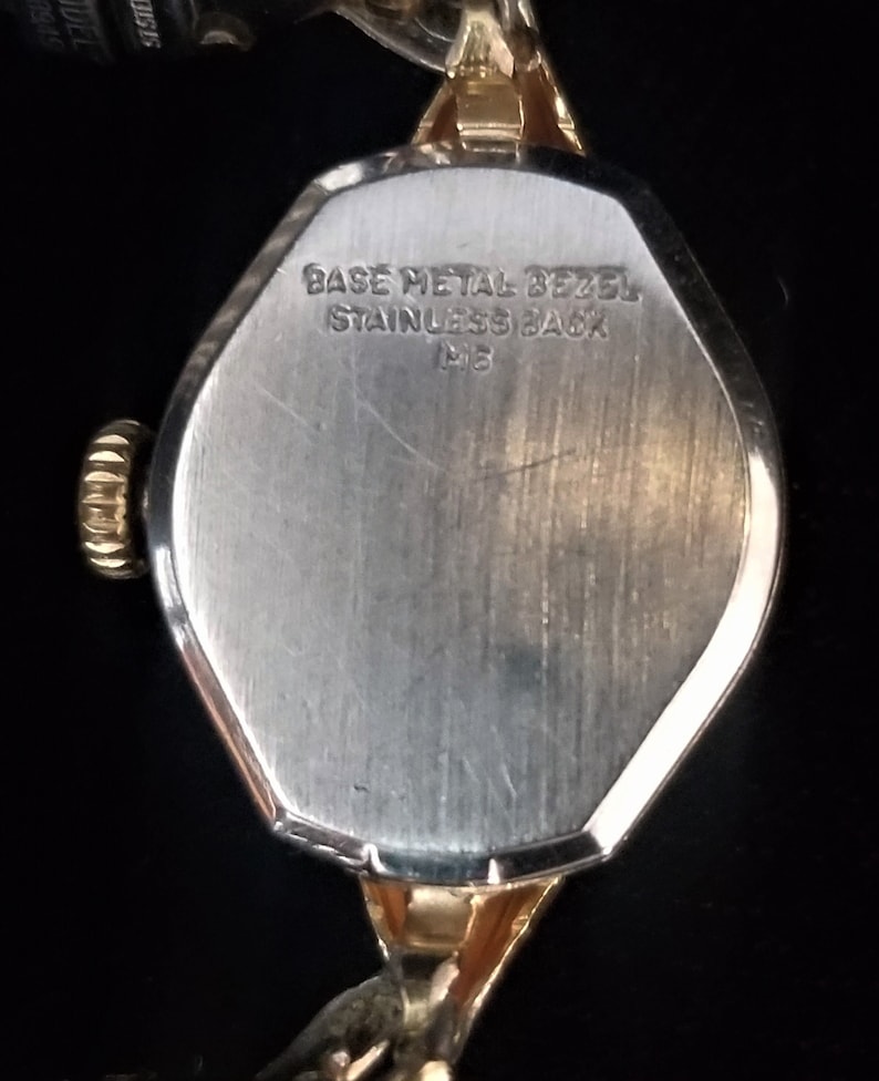 K&ouml;nnte beinhalten: Die R&uuml;ckseite einer goldfarbenen Armbanduhr mit einem silberfarbenen Geh&auml;use. Das Geh&auml;use hat einen erh&ouml;hten Rand und der Text "BASE METAL BEZEL STAINLESS BACK M15" ist in das Geh&auml;use graviert.