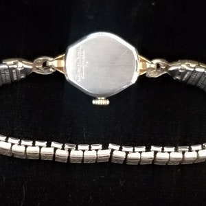 K&ouml;nnte beinhalten: Eine silberne und goldene Armbanduhr mit einem silbernen Armband. Das Zifferblatt der Uhr ist achteckig und hat einen silbernen R&uuml;cken.