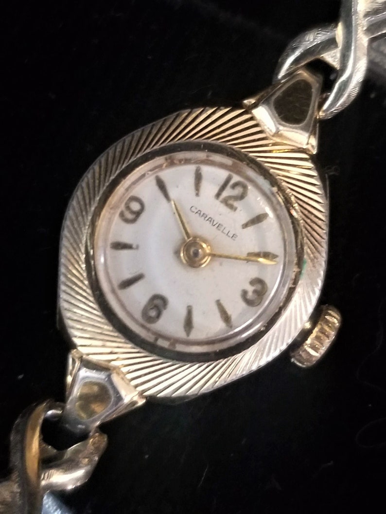 K&ouml;nnte beinhalten: Eine goldfarbene Armbanduhr mit wei&szlig;em Zifferblatt und schwarzen r&ouml;mischen Ziffern. Die Uhr hat ein goldfarbenes Armband mit einem strukturierten Design. Das Zifferblatt der Uhr zeigt "Caravelle".