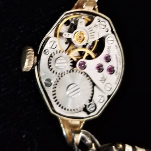K&ouml;nnte beinhalten: Eine goldfarbene Armbanduhr mit silbernem Zifferblatt und sichtbarem mechanischem Uhrwerk. Die Uhr hat eine einzigartige achteckige Form und ein Gliederarmband.