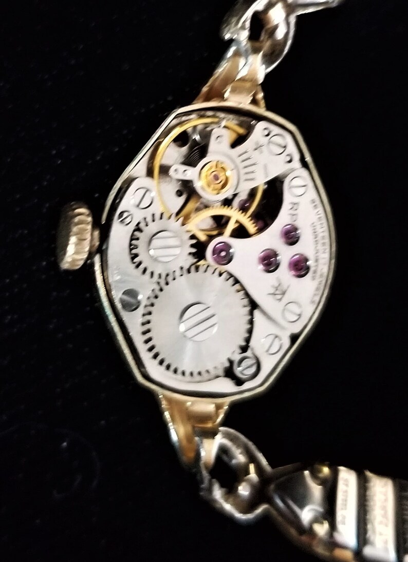 K&ouml;nnte beinhalten: Eine goldfarbene Armbanduhr mit silbernem Zifferblatt und sichtbarem mechanischem Uhrwerk. Die Uhr hat eine einzigartige achteckige Form und ein Gliederarmband.