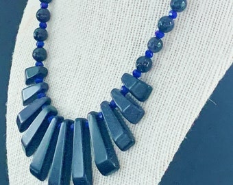 Puede incluir: Un collar negro con un colgante en forma de abanico hecho de piedras negras. El collar está hecho de pequeñas cuentas redondas negras con acentos azules.