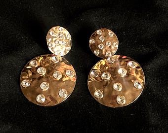 May include: A pair of gold tone earrings with two round discs. The larger disc is textured and has a scattered pattern of small clear rhinestones. The smaller disc is smooth and also has a scattered pattern of small clear rhinestones.
