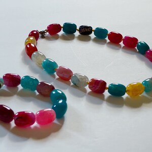 Pu&ograve; includere: Un set di due collane di perline di agata colorate. La collana pi&ugrave; grande &egrave; una lunga fila di perline ovali in tonalit&agrave; di blu, verde, rosso, giallo e rosa. La collana pi&ugrave; piccola &egrave; un braccialetto con perline simili in una lunghezza pi&ugrave; corta.