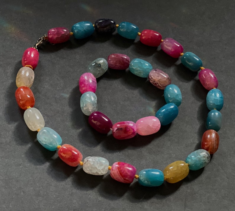 Pu&ograve; includere: Un set di due collane in pietra di agata con una miscela di colori rosa, blu e verde. Le collane sono fatte di perline ovali con accenti dorati.